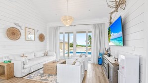 A spacious living room with a white sofa, a coffee table, and a TV mounted on the wall. There is a large window with a view of a beach and a pool. - La Dolce Vita Cottage, Beachfront w/ pool directly on Tropic of Cancer Beach! (Moore Hill)