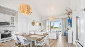 A modern kitchen with white cabinetry, a dining area with wicker chairs, and a large window overlooking a pool. - La Dolce Vita Cottage, Beachfront w/ pool directly on Tropic of Cancer Beach! (Moore Hill)