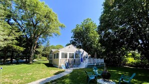 Une petite maison dotée d’un porche, entourée d’arbres et d’une pelouse aménagée avec des sièges d’extérieur.