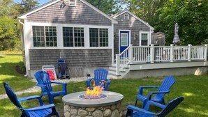 Un jardin avec un foyer en pierre entouré de chaises Adirondack bleues, une terrasse en bois et une maison avec une porte bleue.