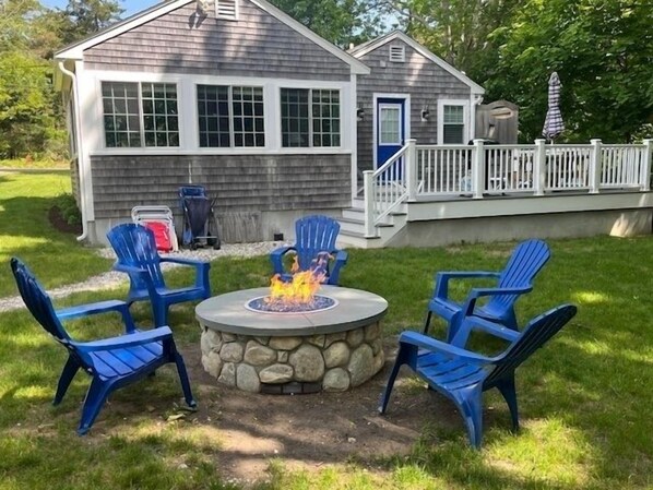 A backyard with a stone fire pit surrounded by blue Adirondack chairs, a wooden deck, and a house with a blue door.