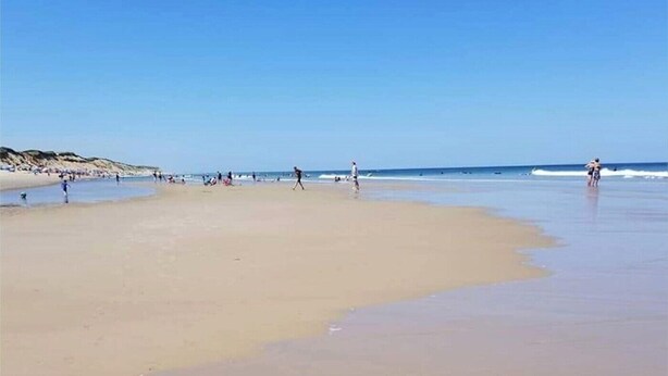 Une vaste plage de sable fin, avec des vagues douces, un ciel dégagé et des collines qui se dessinent à l’horizon.