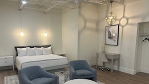 Un dormitorio moderno con una cama grande, dos sillones, un otoman redonda, un escritorio y una lámpara colgante.