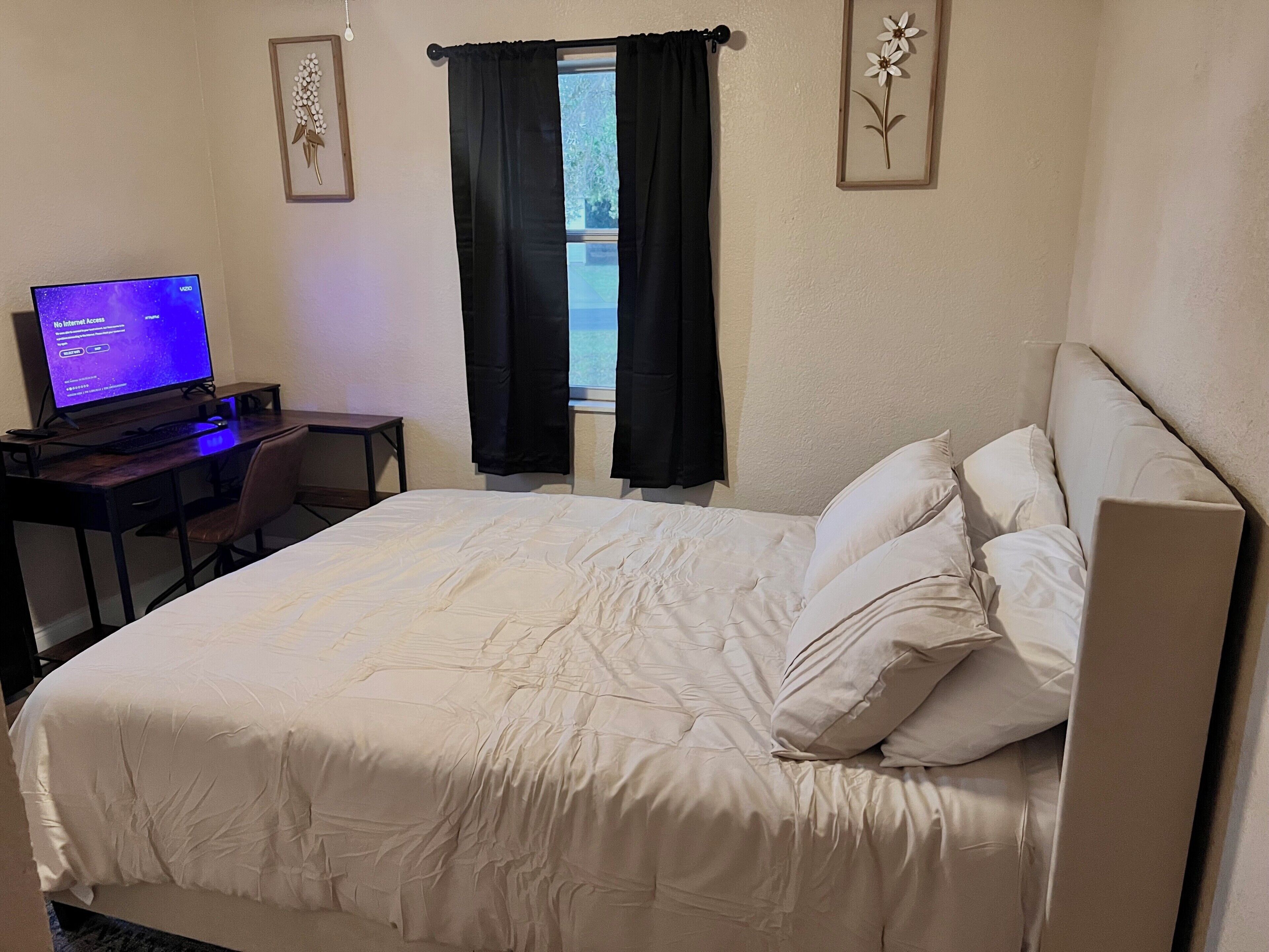 A hotel room with a bed, a desk with a computer, and a window with black curtains.