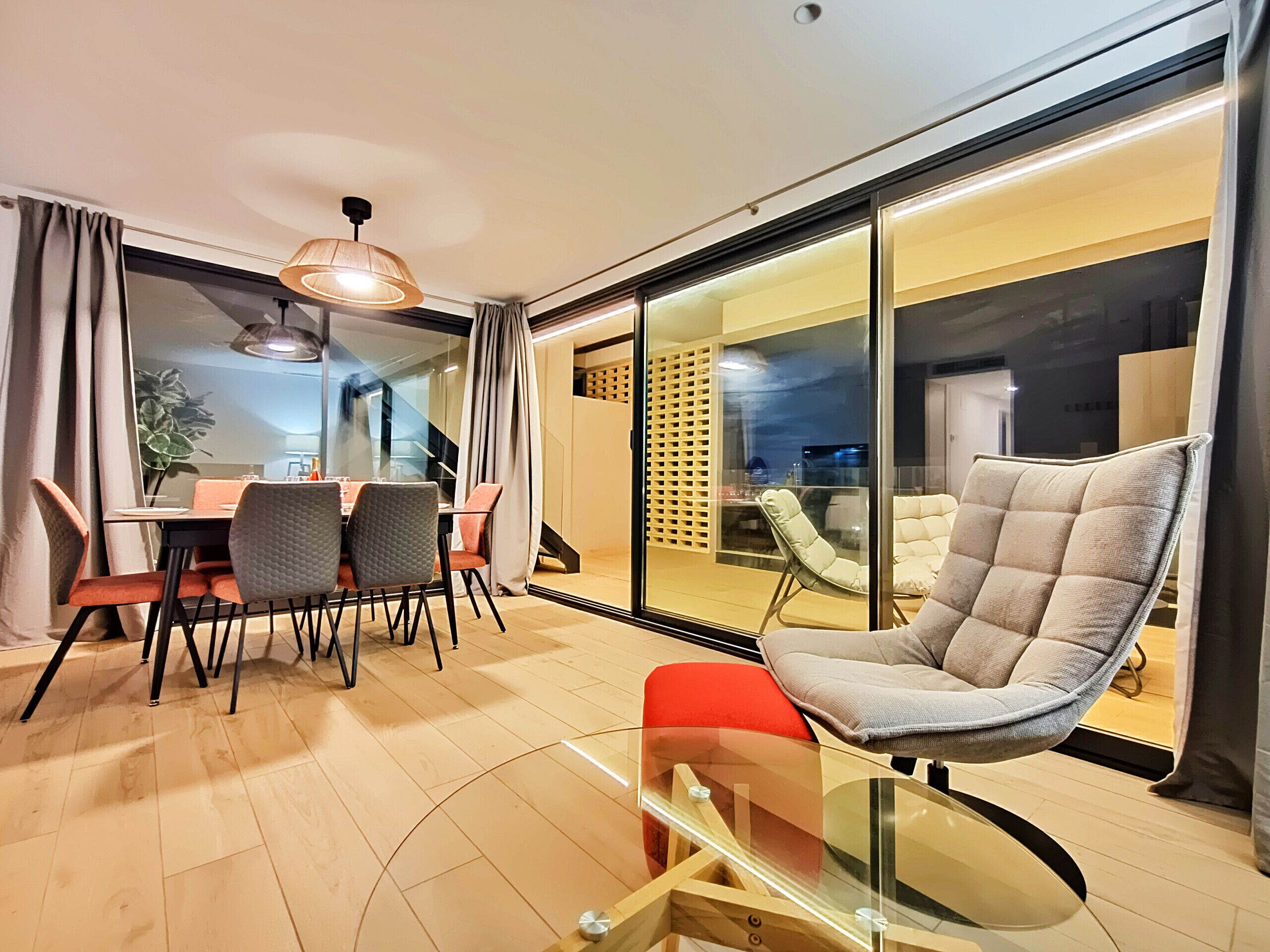 A modern living room with a glass table, a cushioned chair, and a dining area with chairs and a table.