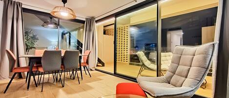 A modern living room with a glass table, a cushioned chair, and a dining area with chairs and a table.
