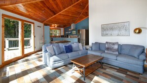 Ein geräumiges Wohnzimmer mit Holzbalkendecke, einem großen, winkelförmigen Sofa und einem Essbereich, der durch eine Glastür sichtbar ist.