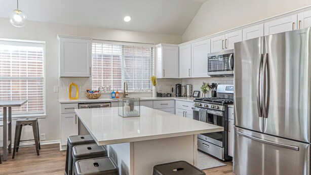 A modern kitchen with white cabinetry, a stainless steel refrigerator, and a central island with stools. - Upscale Home w/ Pool, Arcade & More! (Frisco)