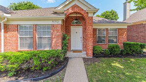 A brick house with a white front door, a number "9712" above the door, and a small garden with bushes and a pathway leading to the door. - Upscale Home w/ Pool, Arcade & More! (Frisco)