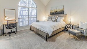 A bedroom with a large bed, a desk with a chair, a side table, and a window with blinds. - Upscale Home w/ Pool, Arcade & More! (Frisco)