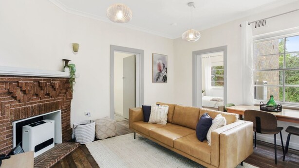 A modern living room with a brick fireplace, a beige sofa, and a large window offering a view of greenery. - Katoomba Getaway 3 bedrooms (Katoomba)