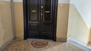 A black double door with a brass handle and a brass lock, a doormat with "WELCOME" written on it, and a beige and white wall with a white electrical outlet.
