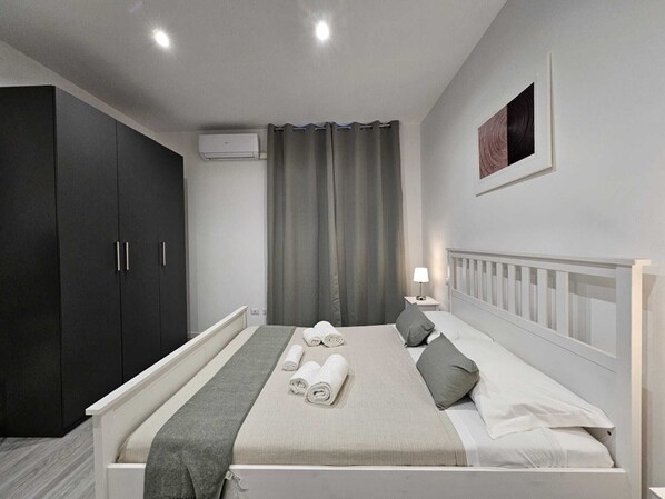 A bedroom with a bed, grey curtains, a white bedside table with a lamp, and a dark wardrobe.