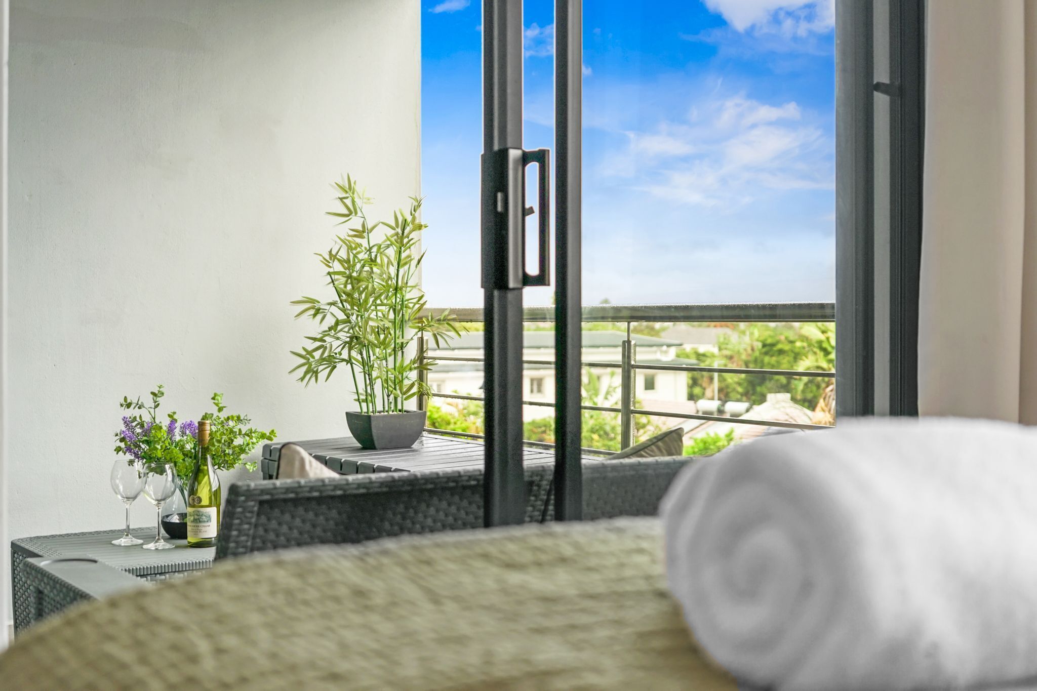 A balcony with a view of buildings, a potted plant, a bottle of wine, and a rolled towel.
