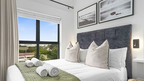 A neatly made bed with white linens and a green throw, a large window with curtains, and a view of greenery outside.