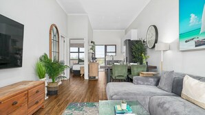 A modern living room with a grey sofa, wooden floor, a coffee table with books, a TV, and a large window.