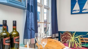 A dining area with two wine bottles, two glasses, a wooden table, a blue curtain, a window with a view, and a sailboat painting on the wall. - Captain House BnB in Fano (Fano)