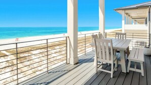 Een overdekt buitengebied met uitzicht op het strand, met een tafel, stoelen en een leuning langs de rand.