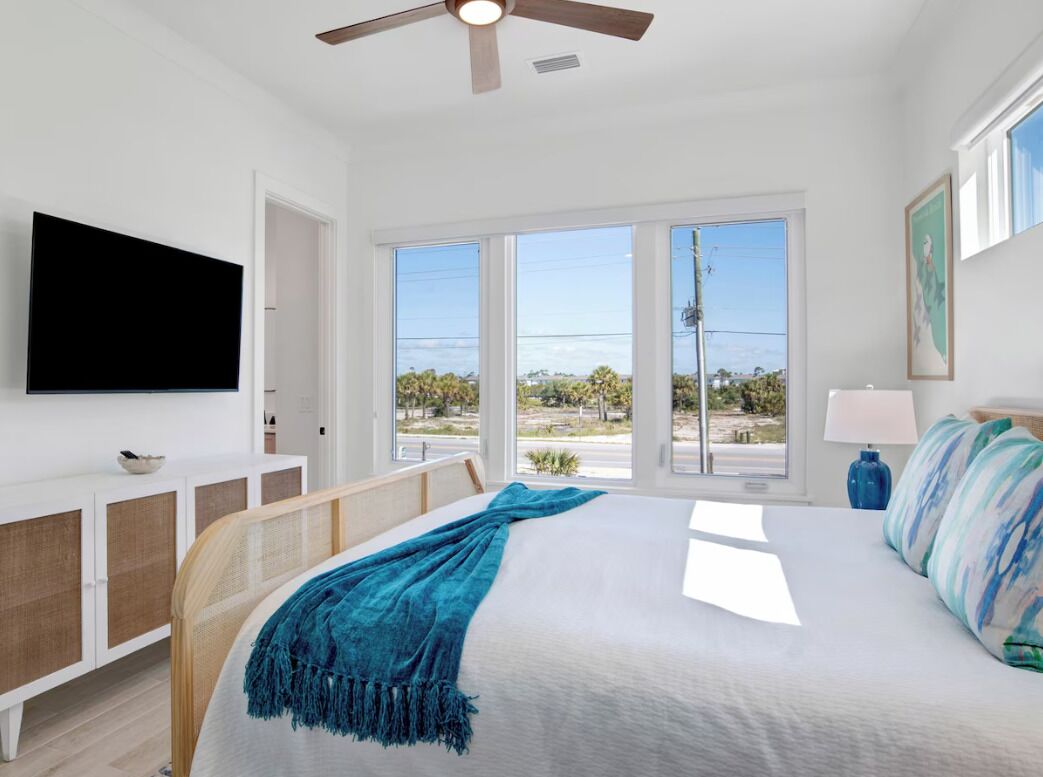 A bedroom with a bed, a ceiling fan, a TV, a window with a view, and a bedside table with a lamp.