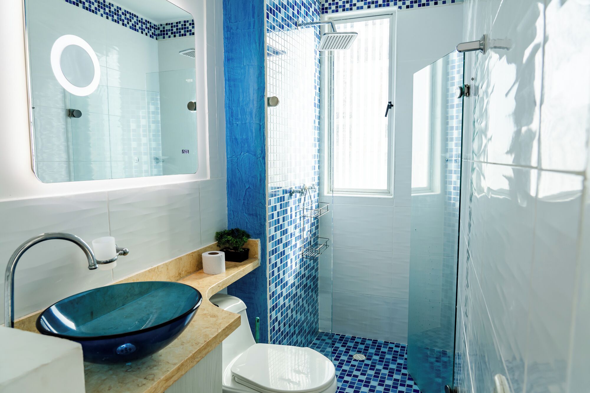 A bathroom with a blue-tiled shower, a white sink with a blue bowl, and a mirror with a circular light.