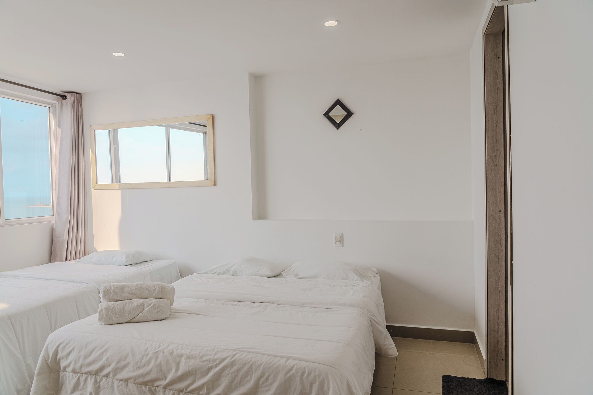 A hotel room with two beds, a window, a mirror, and a light switch on the wall.