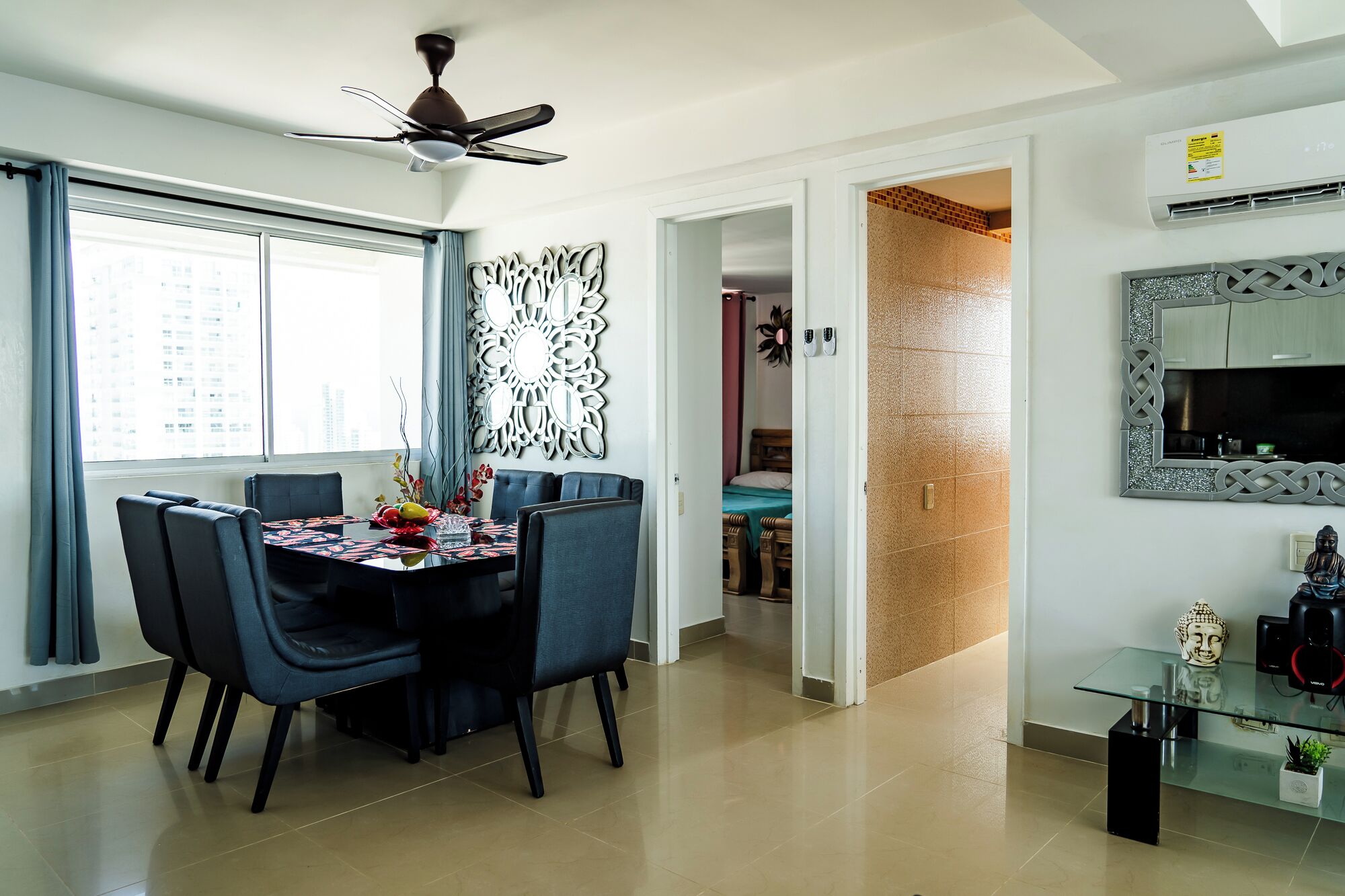 A modern dining area with a round table, chairs, and a ceiling fan. There is a doorway leading to a bedroom with a bed and a bathroom visible.