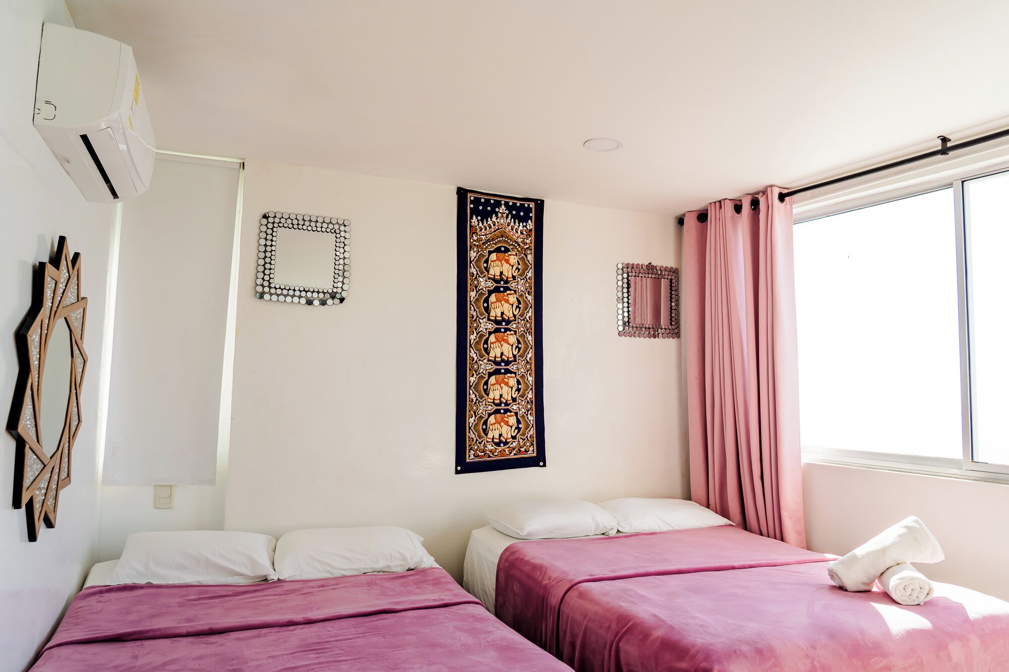 Un dormitorio con dos camas, un ventanal con cortinas rosadas y un aire acondicionado de pared.