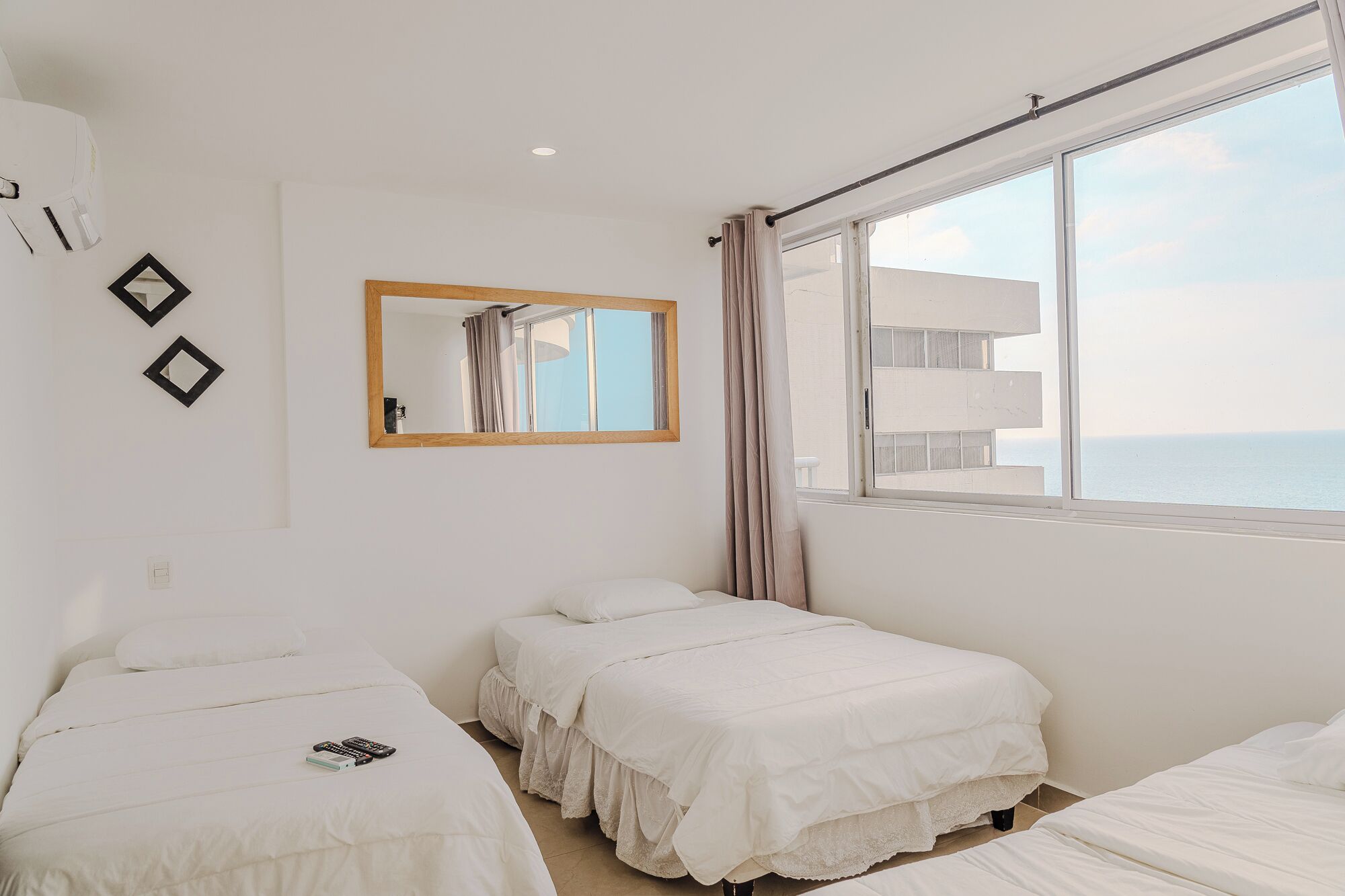 Habitación de hotel con dos camas individuales, un ventanal con vista al mar, un espejo en la pared y una lámpara en el techo.