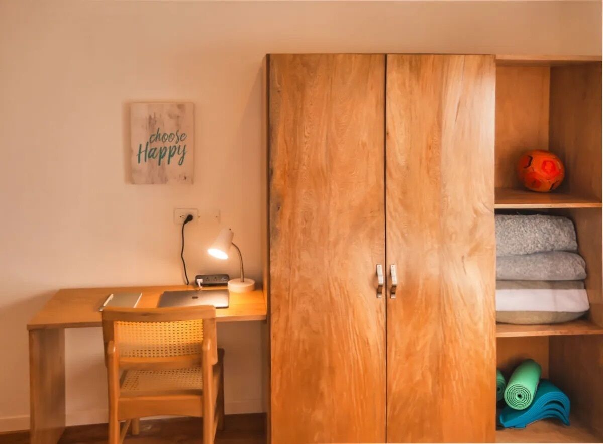 Un bureau en bois avec un ordinateur portable, une lampe et une chaise en osier. On y voit une armoire sur laquelle est accroché un panneau indiquant « Choisissez le bonheur », ainsi qu’une étagère avec des couvertures pliées.