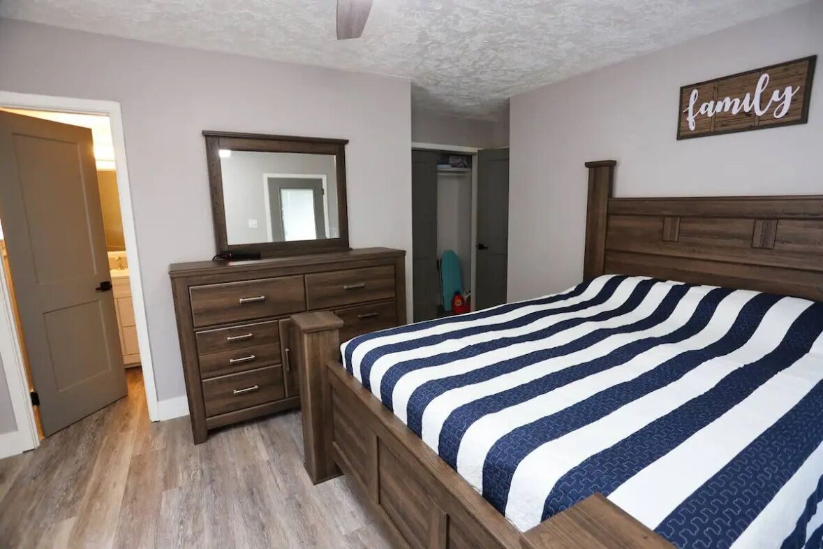 A bedroom with a wooden dresser, a bed with a striped bedspread, a mirror above the dresser, and a 'family' sign on the wall.