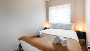 Una cama bien tendida con una manta marrón y dos toallas enrolladas, una mesita de noche con una lámpara, y una ventana con persianas.