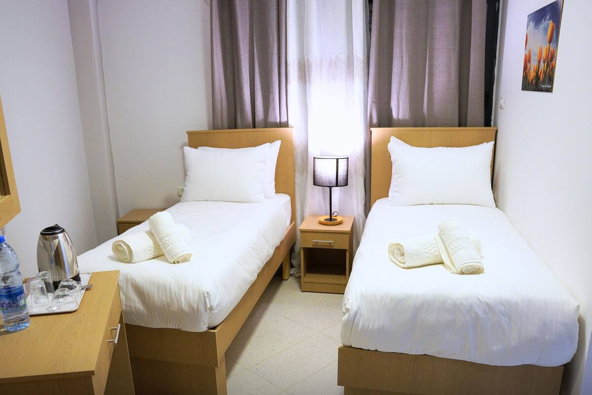 A hotel room with two single beds, a wooden desk with a water bottle, a kettle, and a lamp.