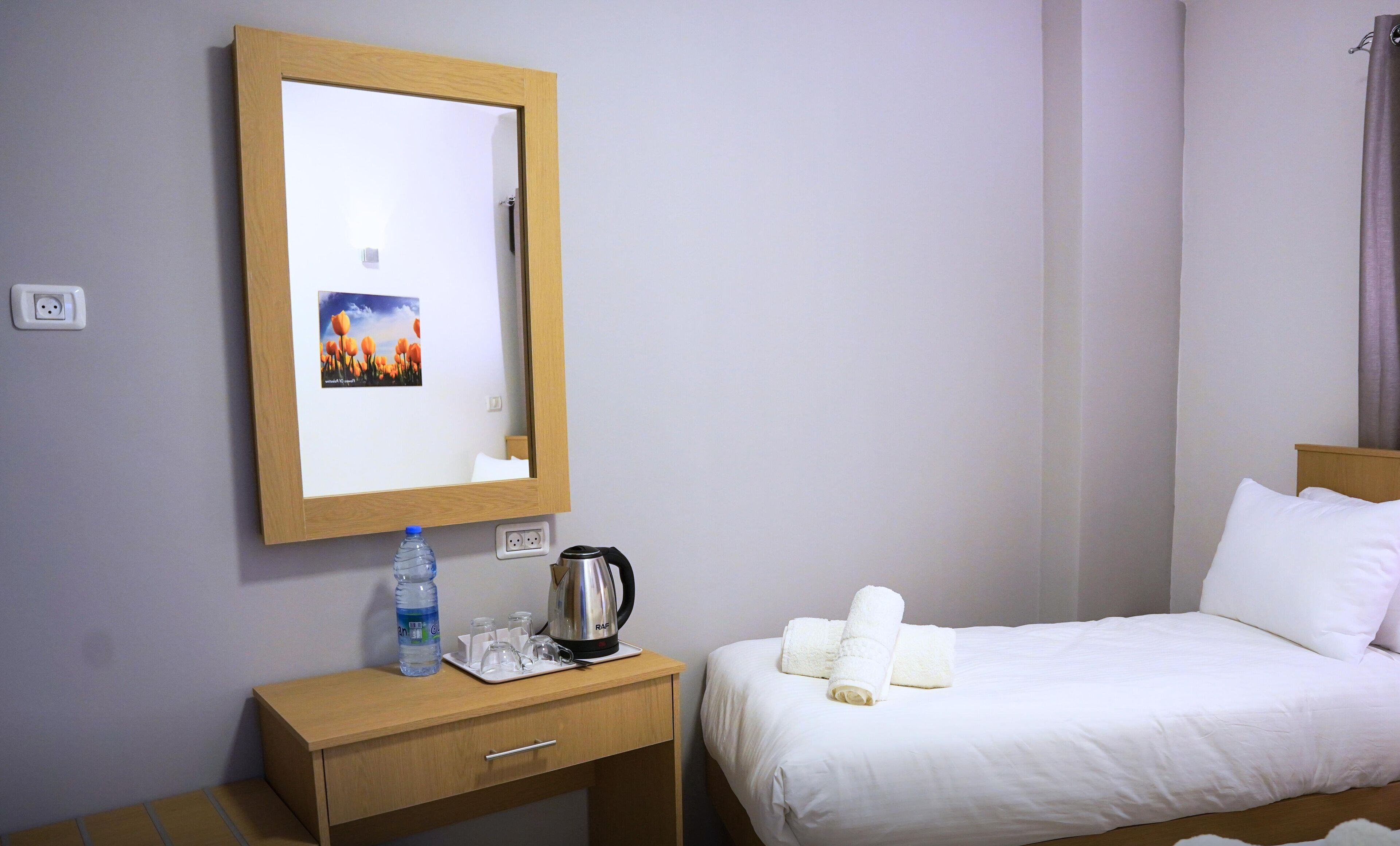 A hotel room with a wooden desk, a bed with white linens, a kettle, a water bottle, and a framed picture on the wall.
