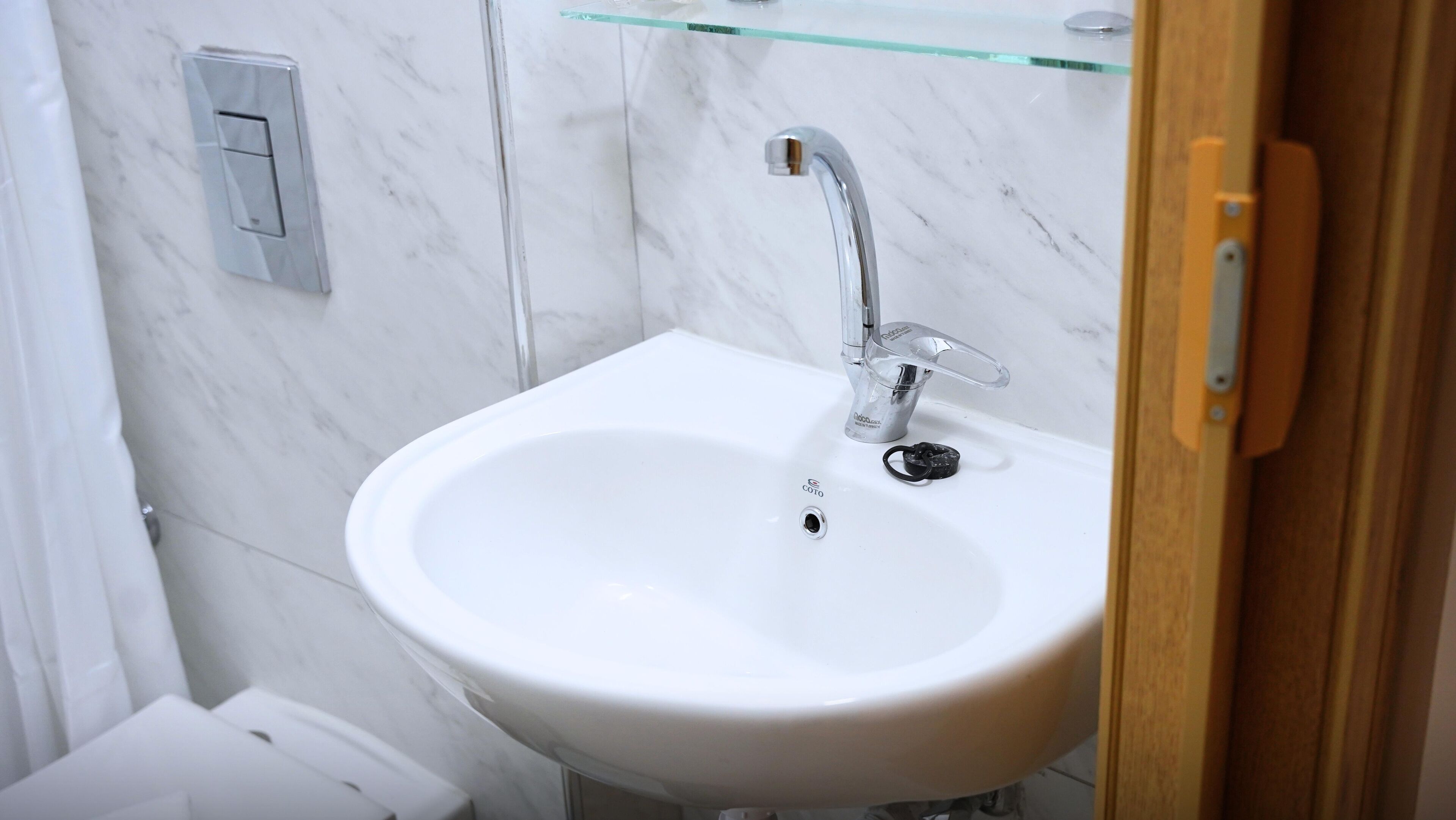 A white bathroom sink with a chrome faucet, a glass shelf above it, and a marble wall.