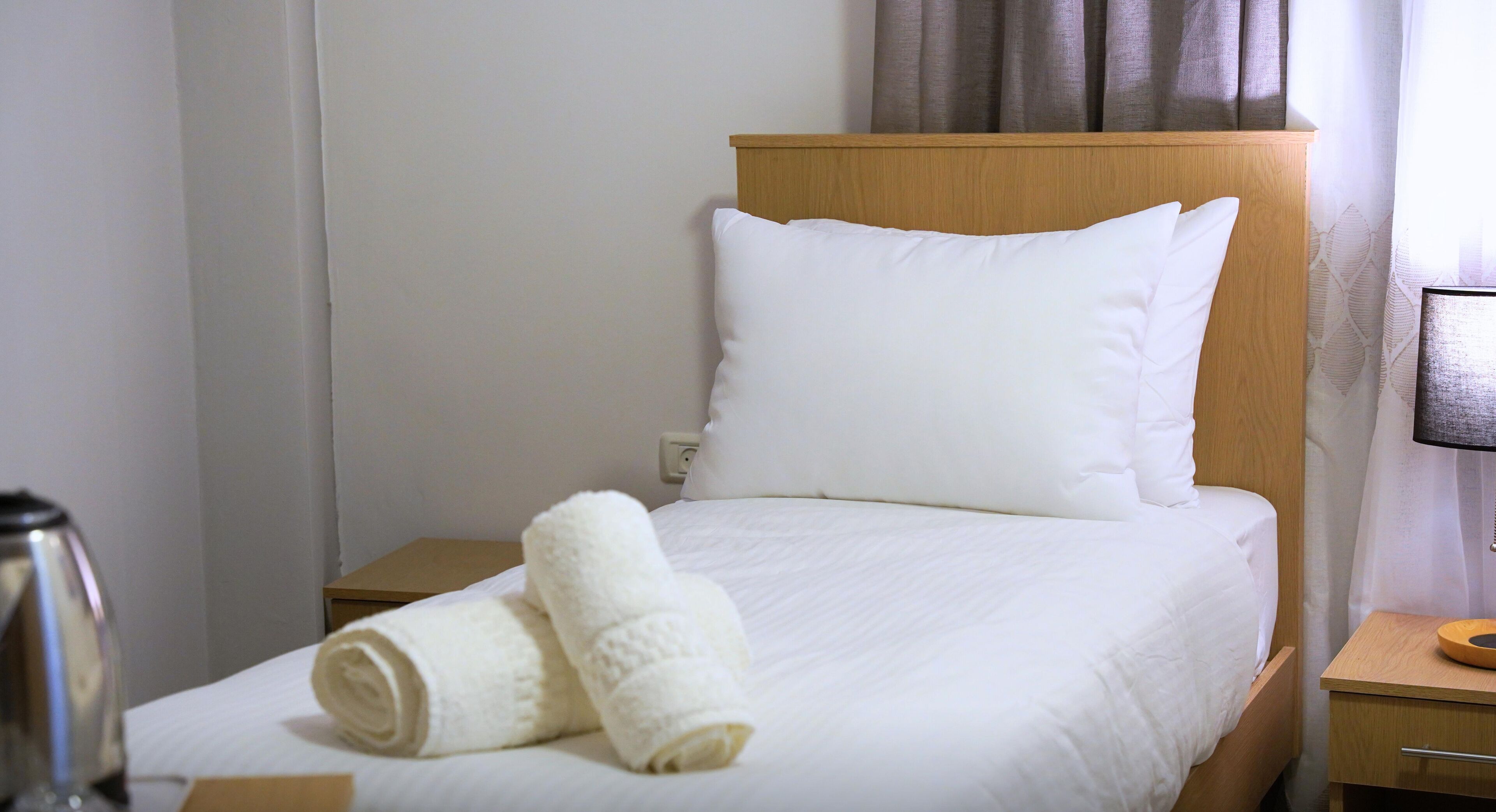A neatly made bed with white linens and two rolled towels, a wooden headboard, a bedside table with a lamp, and a wall socket.