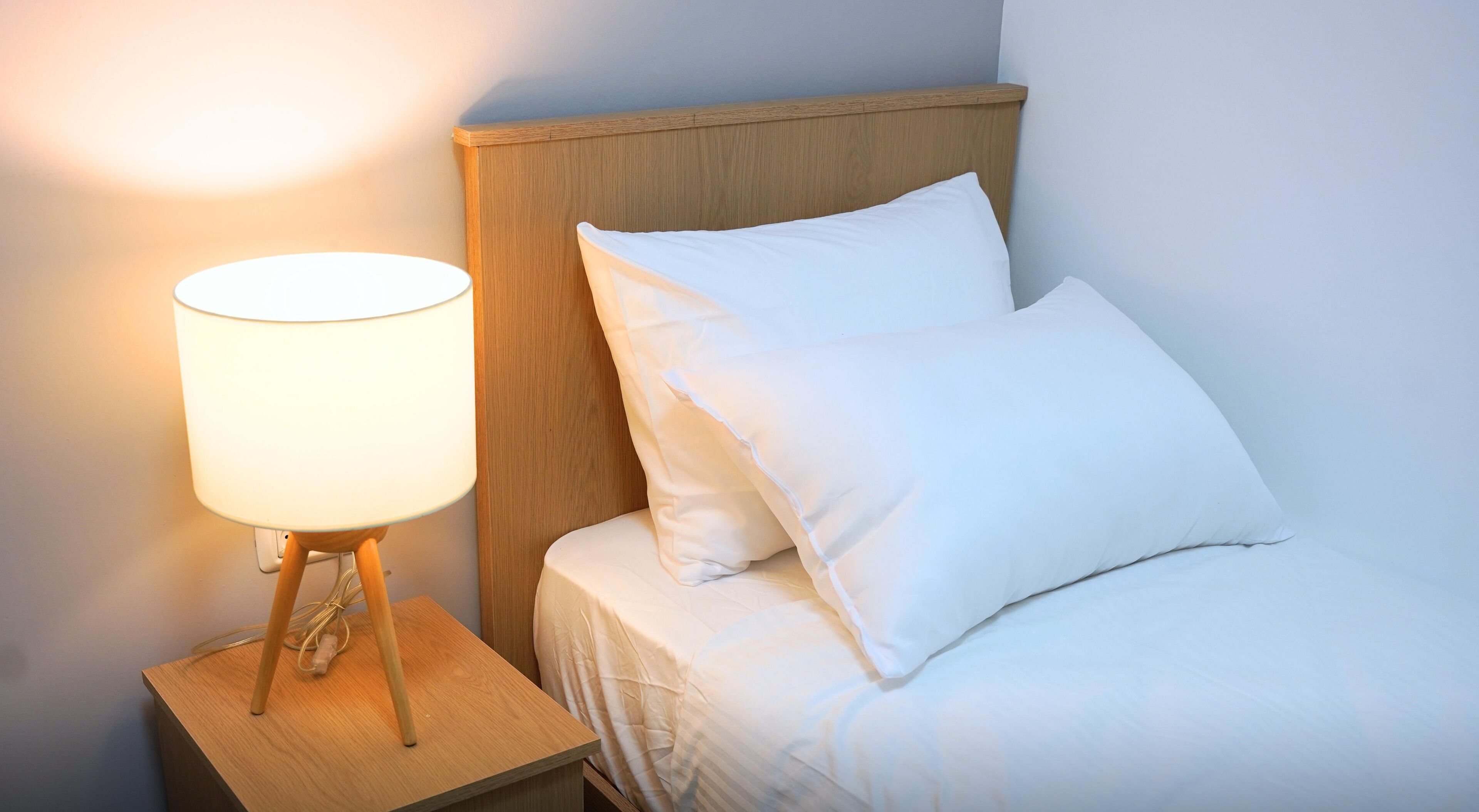 A neatly made bed with white bedding and pillows, a wooden nightstand with a white lamp, and a wooden headboard.
