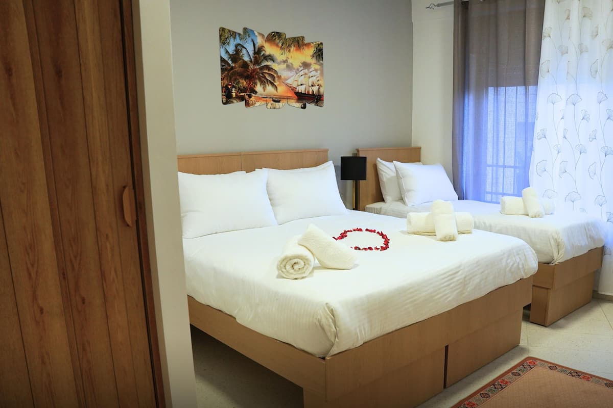 A double room with two beds, white bedding, a wooden headboard, a small bedside table with a lamp, and a tropical artwork on the wall.