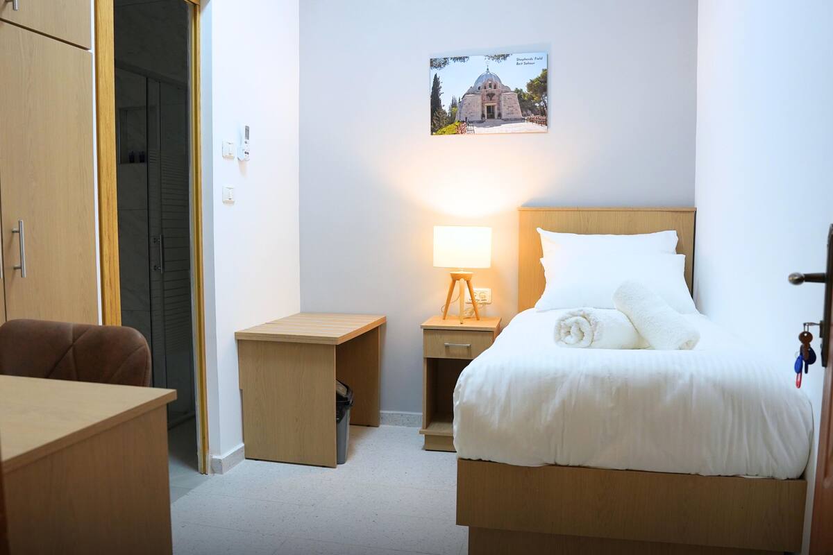 A hotel room with a bed, a desk, a chair, a nightstand with a lamp, and a picture on the wall.