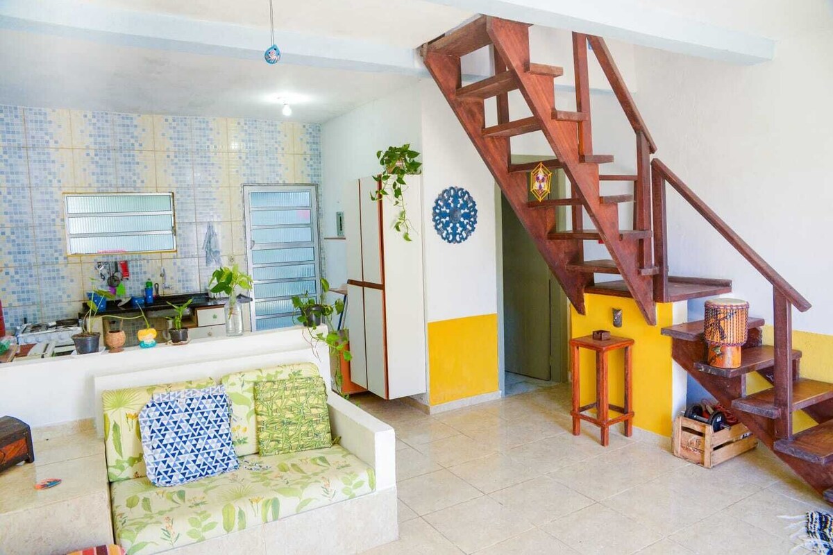 Ein Wohnzimmer mit einer Holztreppe, einem Sofa mit gemusterten Kissen und einer Küchenzeile mit gefliestem Fliesenspiegel und Blumentöpfen.