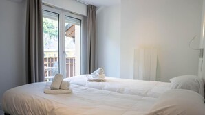 A neatly made bed with white linens and towels, a large window with curtains, and a view of buildings outside. - Wifi, resort center, fitness, balcony, tv, 42m², Brides Les Bains (Brides-les-Bains)