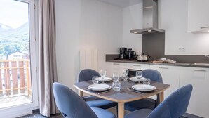 A dining area with a table set for four, a kitchen in the background, and a view of a mountainous landscape through a large window. - Wifi, resort center, fitness, balcony, tv, 42m², Brides Les Bains (Brides-les-Bains)