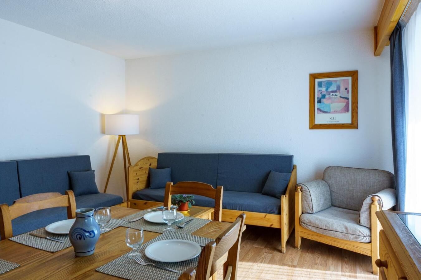 Un comedor con una mesa de madera dispuesta para dos personas, dos sofás azules, un sillón de madera y una lámpara de pie.