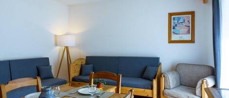 Un comedor con una mesa de madera dispuesta para dos personas, dos sofás azules, un sillón de madera y una lámpara de pie.