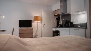 A modern hotel room with a bed, a TV, a desk, a lamp, and a kitchenette.
