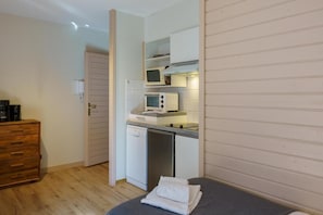 Kompakti keittiö, jossa on mikroaaltouuni, leivänpaahdin ja astianpesukone. Lisäksi näkyvissä on puinen lipasto ja sängyllä oleva taitettu pyyhe.