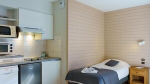 Een compacte hotelkamer met kitchenette, een bed met kussen, een houten nachtkastje en een kleine houten stoel.