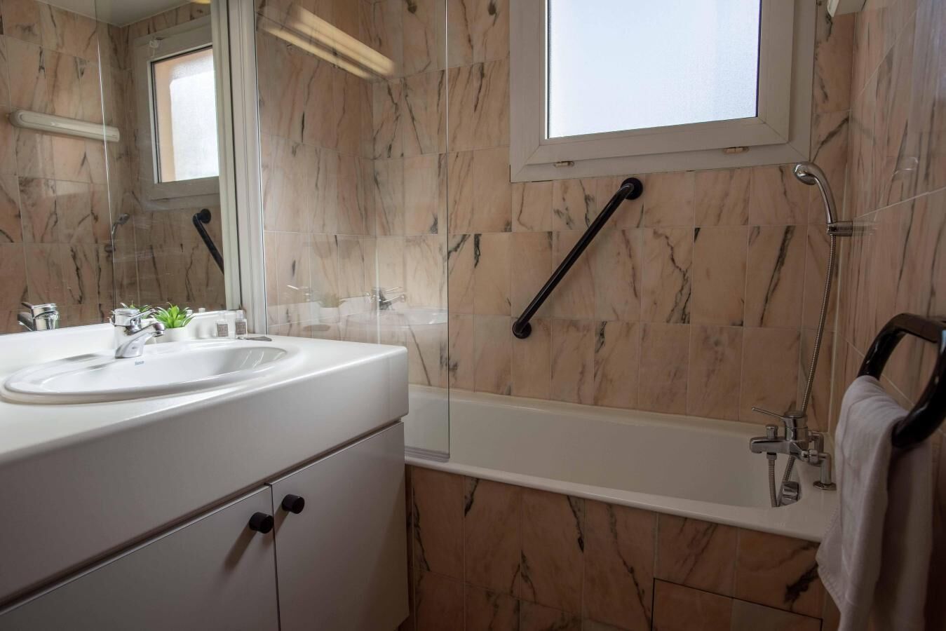 Ein Badezimmer mit einem weißen Waschbecken, einem Spiegel und einer Badewanne. Über dem Spiegel befindet sich ein Fenster und rechts neben der Badewanne ein Handtuchhalter.
