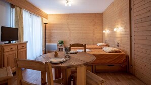 Acogedora habitación tipo cabaña con detalles de madera, una mesa de comedor con sillas, una cama con almohadas, un televisor y una pequeña mesa con una planta.