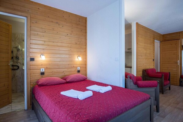 A room with a wooden paneled wall, a bed with red bedding, two bedside lamps, and a chair with red cushions.