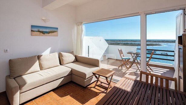 A modern living room with a beige sectional sofa, a small wooden table, and a view of the sea through large glass doors.